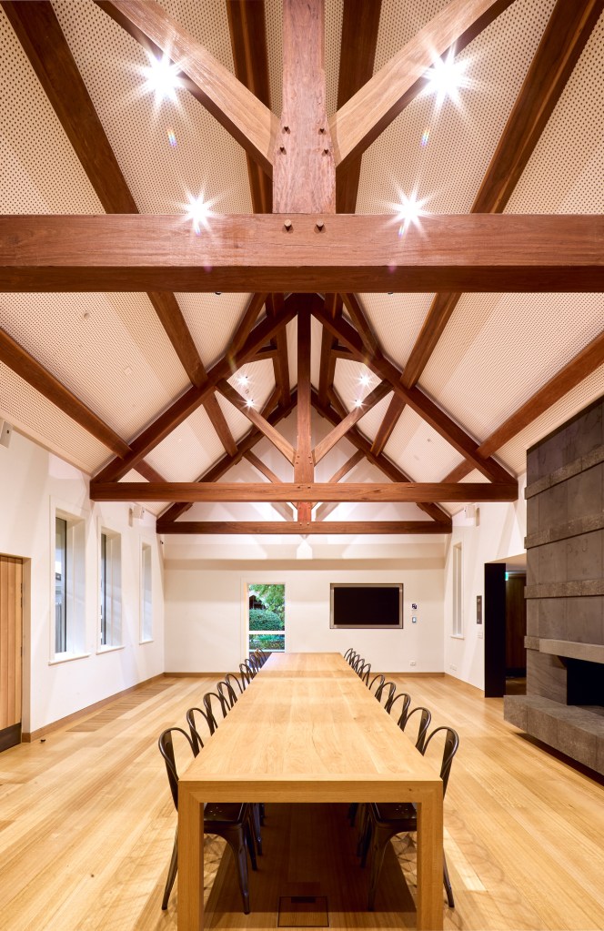 A large room with a high, open ceiling and a long table surrounded by chairs. The ceiling has wooden trusses and a white f...